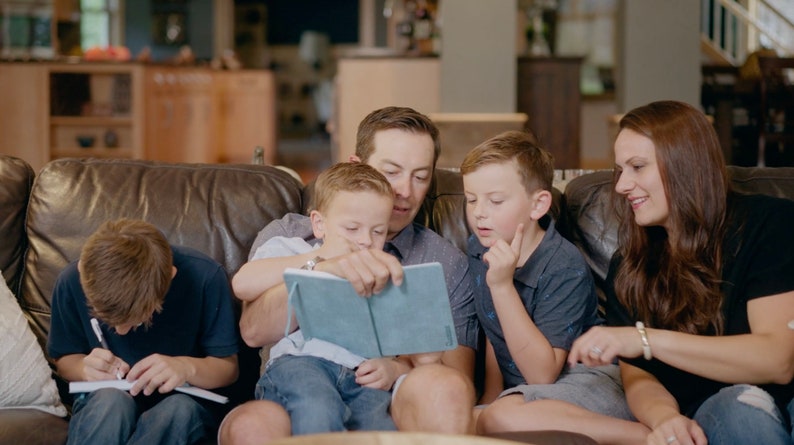 Puede incluir: Una familia de cuatro personas sentada en un sof&aacute; de cuero marr&oacute;n. Un hombre est&aacute; leyendo un libro azul a dos ni&ntilde;os. Una mujer est&aacute; sentada junto a los ni&ntilde;os y sonriendo. El ni&ntilde;o de la izquierda est&aacute; escribiendo en un cuaderno.