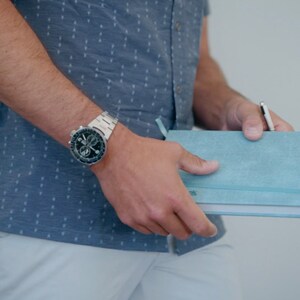 Puede incluir: Una persona con una camisa azul con estampado y pantalones blancos sostiene un cuaderno azul claro con un bol&iacute;grafo que sobresale. La persona lleva un reloj plateado con una esfera negra.