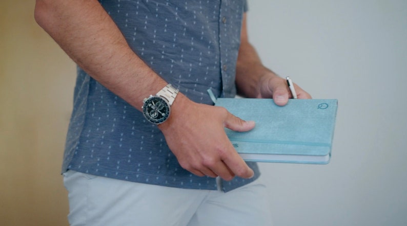 Puede incluir: Una persona con una camisa azul con estampado y pantalones blancos sostiene un cuaderno azul claro con un bol&iacute;grafo que sobresale. La persona lleva un reloj plateado con una esfera negra.