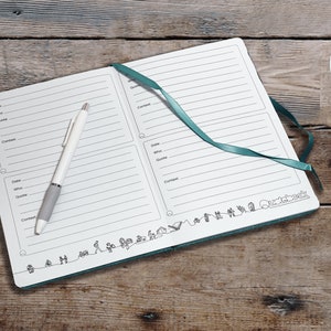 Puede incluir: Un cuaderno blanco con una cubierta turquesa y un bol&iacute;grafo encima. El cuaderno tiene p&aacute;ginas rayadas y una secci&oacute;n para notas. La cubierta tiene un borde decorativo con peque&ntilde;os dibujos de personas y &aacute;rboles. El cuaderno est&aacute; sobre una superficie de madera.