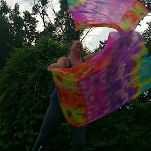 May include: A person is holding and twirling two large, colorful fabric flags. The flags are tie-dyed with shades of blue, pink, purple, orange, and yellow. The person is wearing dark pants and a blue tank top.