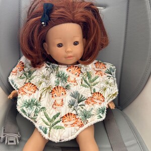 Könnte beinhalten: Eine Puppe mit roten Haaren und einer schwarzen Schleife, die ein Lätzchen mit einem Tiger- und Blättermuster trägt. Die Puppe sitzt in einem grauen Kinderwagen. Das Lätzchen zeigt orange und braune Tiger mit grünen Blättern auf weißem Grund.