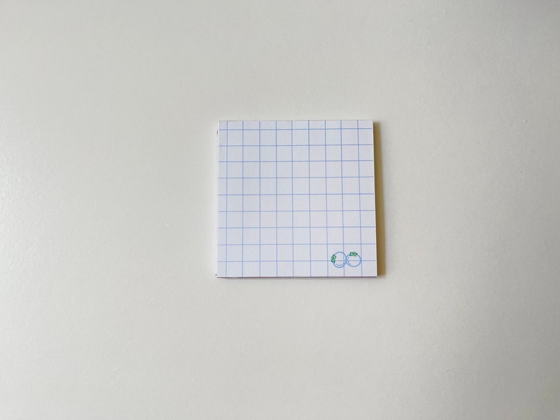 May include: A white notepad with blue grid lines and a small green drawing of two blueberries on the bottom right corner.
