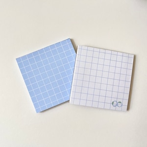 May include: Two square sticky note pads with a grid pattern. One pad is light blue with white grid lines, the other is white with light blue grid lines. The white pad has a small green design in the bottom right corner.