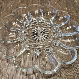 May include: A clear glass egg serving platter featuring a circular design with twelve individual oval-shaped indentations arranged around a central starburst pattern. The plate has a scalloped edge and a smooth, reflective surface throughout.