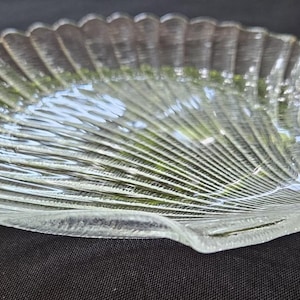 以下が含まれることがあります： テクスチャ加工された表面を持つ、透明なガラス製の貝殻型皿。 皿はスカラップエッジと湾曲した扇のようなデザインです。 この皿は約25cmの長さと18cmの幅で、軽食の提供や装飾品の展示に最適です。