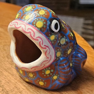 Könnte beinhalten: Eine Keramik-Fischfigur mit offenem Maul. Der Fisch ist mit einer braunen Basis, blauen Akzenten und gelben und rosa Details bemalt. Das offene Maul ist weiß mit rosa Akzenten, und das Auge ist schwarz und weiß.