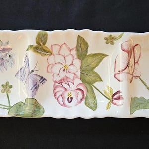 Pode incluir: Uma travessa retangular de cerâmica branca com três compartimentos. A travessa é decorada com flores rosa e roxas pintadas à mão, folhas verdes e uma borboleta. As bordas têm um design ondulado.