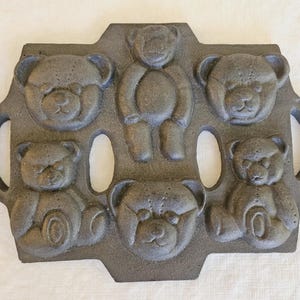 Puede incluir: Un molde para hornear de hierro fundido gris oscuro con seis diseños de osos de peluche. El molde tiene dos asas y está diseñado para hornear pasteles o golosinas. Los osos están en varias poses, incluyendo sentados y de pie. El molde es de hierro fundido.