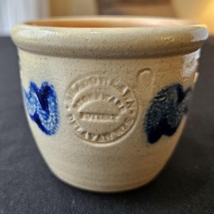 Può includere: Un piccolo vaso in ceramica color crema con un motivo floreale blu e bianco. Il vaso ha una bocca rotonda e larga e un fondo arrotondato. È stampato con le parole "Crocksville, Ohio" e "USA".