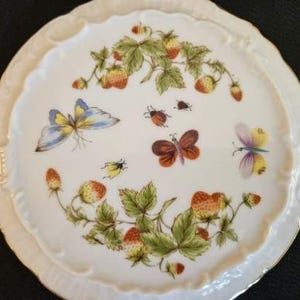 Könnte beinhalten: Ein weißer Keramikteller mit gewelltem Rand, der ein gemaltes Design aus Erdbeeren, grünen Blättern und bunten Schmetterlingen und Insekten zeigt. Der Teller hat einen goldenen Rand.