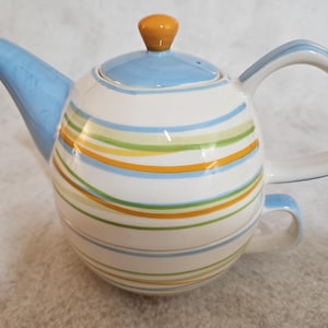 May include: A ceramic teapot with a white base, adorned with horizontal stripes in blue, green, and orange. The spout, handle, and lid are light blue, with an orange knob on top. The teapot has a capacity of approximately 1.2 liters.