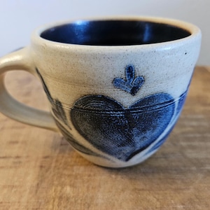 May include: A white ceramic mug with a blue heart design. The mug has a handle and a blue glaze on the inside.