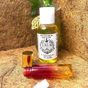 以下が含まれることがあります： 「PURE PERFUME OIL MADE IN USA」と書かれたラベルが付いた、白いキャップの黄色い香水オイルのボトル。小さく空のボトルは赤とオレンジ色です。金色のキャップと白いアプリケーターも表示されています。