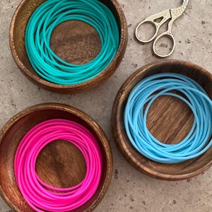 May include: Three round wooden bowls containing coiled silicone cords in bright turquoise, neon pink, and light blue. A pair of silver-toned metal stork-shaped embroidery scissors with intricate handle detailing rests near the bowls.