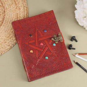 May include: A red leather journal with an embossed pentagram design and colorful gemstone accents. The journal features a metal clasp closure and is surrounded by pencils and small black objects.