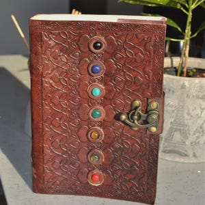 May include: A brown leather journal with embossed floral patterns and a metal clasp. Seven colorful gemstones are vertically aligned on the cover. The journal is closed, suggesting it's ready for writing or sketching.