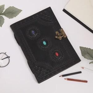 May include: Black leather journal with embossed floral patterns and three embedded gemstones in blue, turquoise, and red. The journal has a brass clasp closure and is surrounded by pencils and leaves.