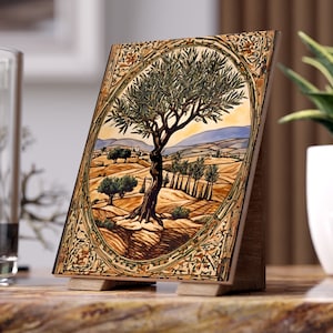 May include: A ceramic tile with a detailed painting of an olive tree in a landscape setting. The tree is brown with green leaves and is surrounded by hills and other trees. The tile is framed with a decorative border.