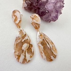 May include: A pair of teardrop-shaped earrings with a marbled design. The earrings feature a mix of white, tan, and gold colors. The earrings are hanging from gold-colored posts. A purple crystal cluster is in the background.