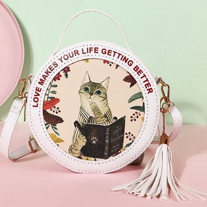 May include: A white round crossbody bag with a red and white print that says 'Love makes your life getting better.' The bag has a white tassel and a cat illustration with a book that says 'The Oxford Sampler'.