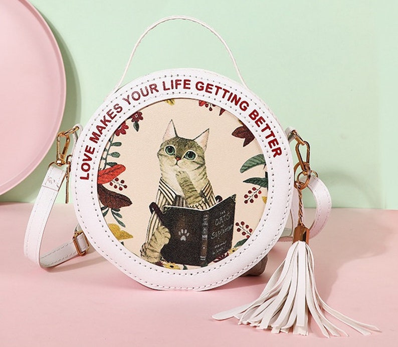 May include: A white round crossbody bag with a red and white print that says 'Love makes your life getting better.' The bag has a white tassel and a cat illustration with a book that says 'The Oxford Sampler'.