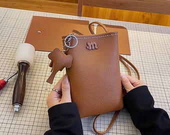 Peut inclure: Un sac à bandoulière pour téléphone en cuir marron avec un petit charme éléphant attaché à la fermeture éclair. Le sac a un détail cousu et la lettre "M" sur le devant.