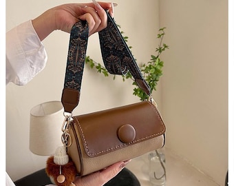 May include: A brown and beige shoulder bag with a patterned strap. The bag has a round button detail and a small, fluffy charm attached.