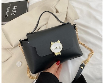 May include: A black faux leather shoulder bag with a gold chain strap and a cute cat face clasp.