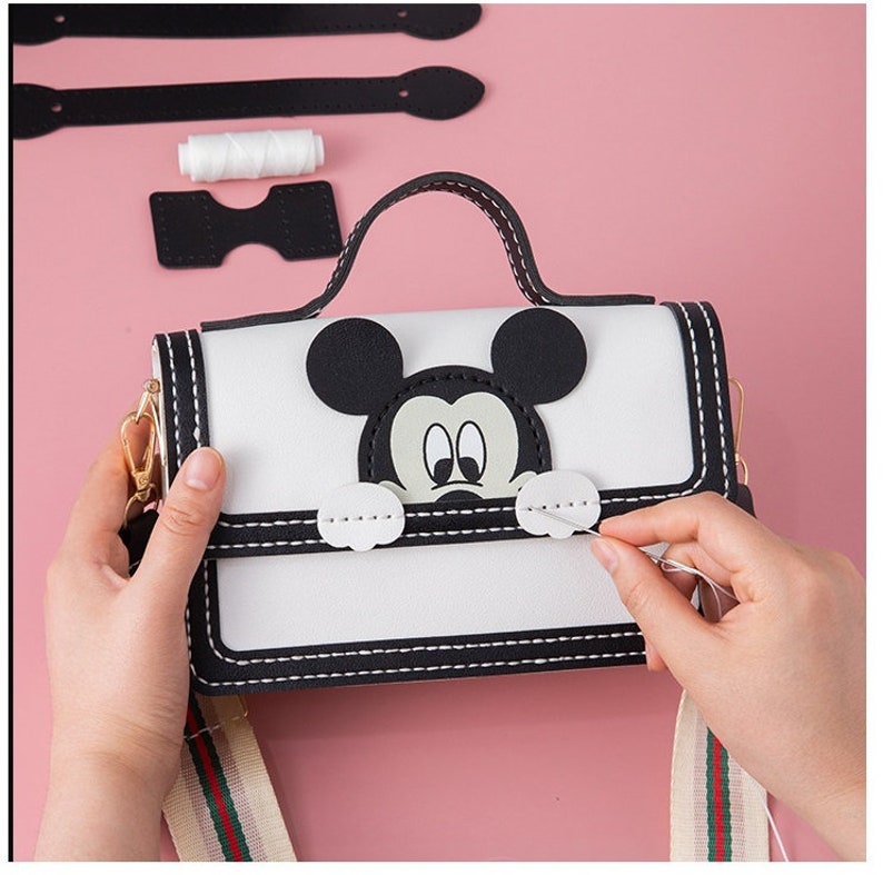 以下が含まれることがあります： ミッキーマウスのキャラクターデザインが施された白黒のフェイクレザーのハンドバッグ。ハンドバッグはトップハンドルと長い調節可能なストラップが付いています。ハンドバッグは白い糸で縫われています。