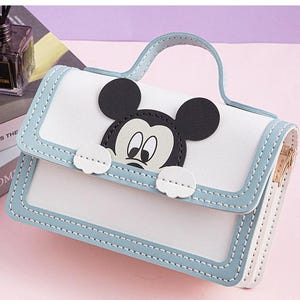 以下が含まれることがあります： ミッキーマウスのキャラクターデザインが施された白と青のハンドバッグ。バッグはジッパーで閉じられ、トップハンドルが付いています。