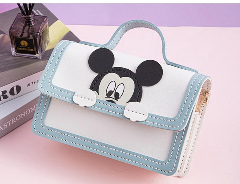以下が含まれることがあります： ミッキーマウスのキャラクターデザインが施された白と青のハンドバッグ。バッグはジッパーで閉じられ、トップハンドルが付いています。
