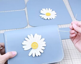 May include: A light blue leather purse with a white daisy flower applique. The purse is being sewn together with white thread.