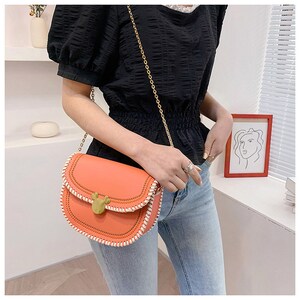 May include: A small orange crossbody bag with a white stitched trim and a gold Mickey Mouse shaped clasp. The bag has a gold chain strap.