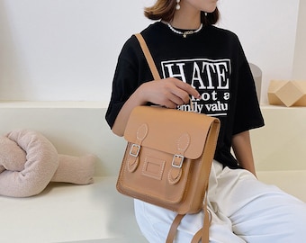 May include: A tan leather satchel-style backpack with two buckles and a front pocket. The bag has a top handle and adjustable shoulder straps. The image also shows a black t-shirt with the word "HATE" printed on it.