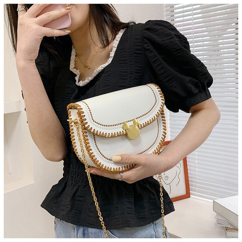 May include: White faux leather shoulder bag with a brown stitched trim and a gold Mickey Mouse shaped clasp. The bag has a gold chain strap.