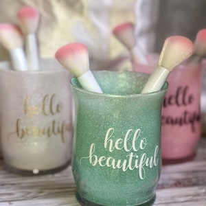 May include: Glitter-covered glass holders in pastel colors, each with the words "hello beautiful" in script. Makeup brushes with white handles and pink tips are inside the holders. The holders are on a light wood surface.