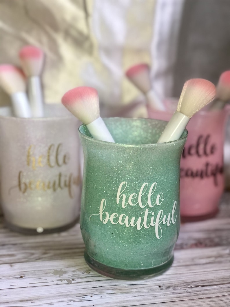 May include: Glitter-covered glass holders in pastel colors, each with the words "hello beautiful" in script. Makeup brushes with white handles and pink tips are inside the holders. The holders are on a light wood surface.