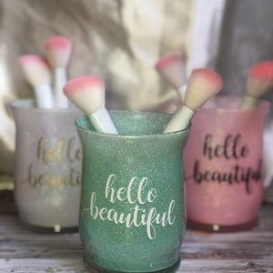 May include: Three glittery makeup brush holders in pastel colors: green, pink, and white. Each holder has the words "hello beautiful" in a cursive font. The holders contain white-handled makeup brushes with pink brush heads.