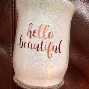 May include: Glitter-covered glass candle holder with the words "hello beautiful" in rose gold script. The candle holder has a wide base and a slightly flared top, perfect for home decor.