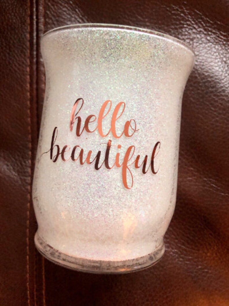 May include: Glitter-covered glass candle holder with the words "hello beautiful" in rose gold script. The candle holder has a wide base and a slightly flared top, perfect for home decor.