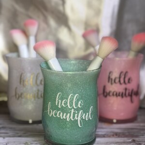 May include: Three glittery makeup brush holders in pastel colors: green, pink, and white. Each holder has the words "hello beautiful" in a cursive font. The holders contain white-handled makeup brushes with pink brush heads.