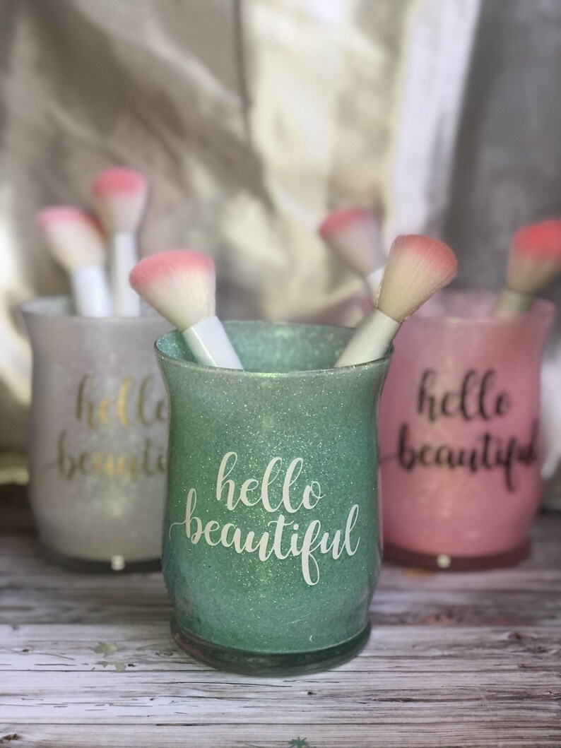 May include: Three glittery makeup brush holders in pastel colors: green, pink, and white. Each holder has the words "hello beautiful" in a cursive font. The holders contain white-handled makeup brushes with pink brush heads.