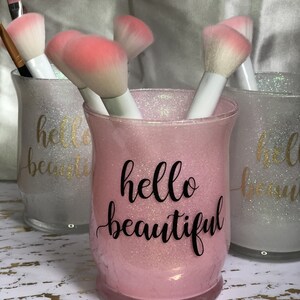 May include: Pink glittery makeup brush holder with the text "hello beautiful" in black script. The holder contains several makeup brushes with white handles and pink and white bristles. Two other similar holders are visible.