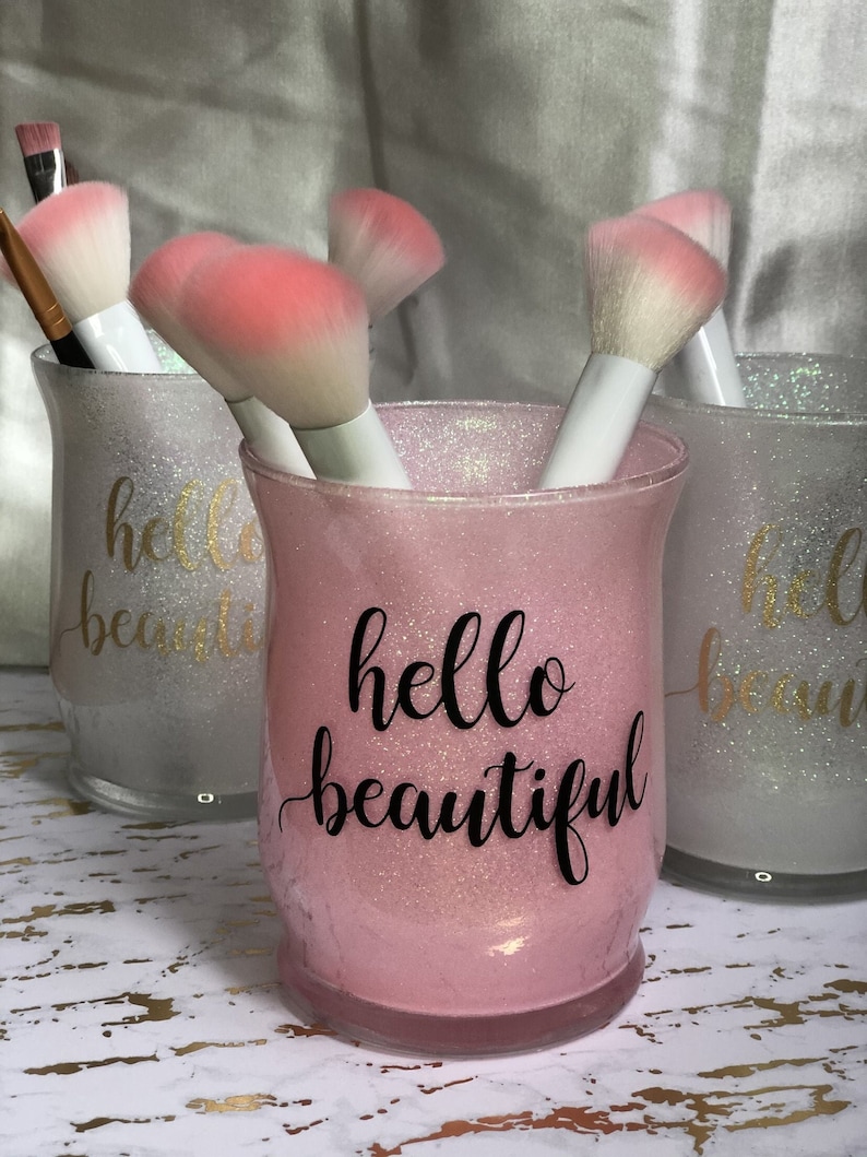 May include: Three decorative glittery glass containers holding makeup brushes. The central container is pink with the words "hello beautiful" in black script. The other two containers are silver with gold script.