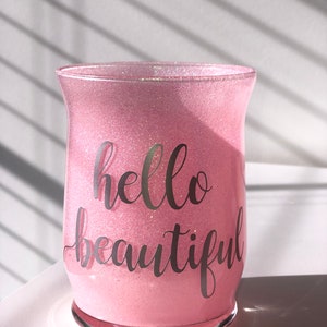 May include: A pink glitter-covered glass candle holder with the words "hello beautiful" in a cursive font. The glass has a flared shape and sits on a clear base. The background is a white surface with shadows.