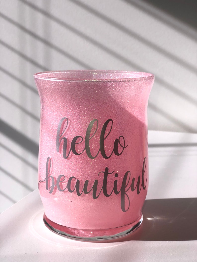 May include: A pink glitter-covered glass candle holder with the words "hello beautiful" in a cursive font. The glass has a flared shape and sits on a clear base. The background is a white surface with shadows.