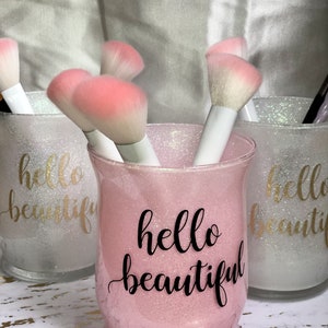 May include: Pink glittery glass jar with the words "hello beautiful" in black script, holding makeup brushes with white handles and pink bristles. Two other jars with the same design are visible in the background. The jars are on a gold and white patterned surface.