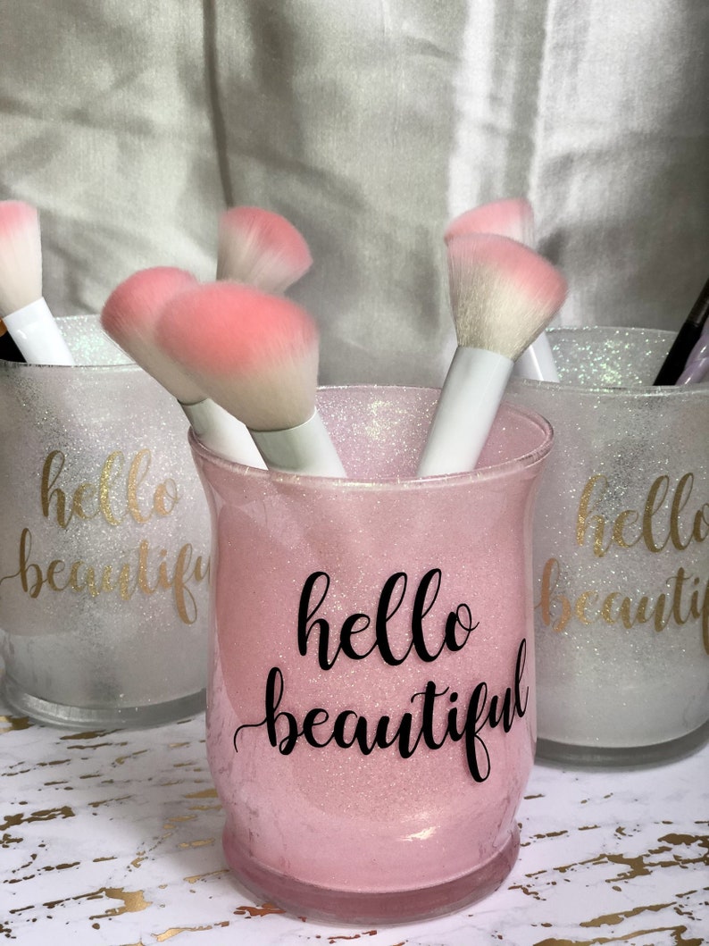 May include: Pink glittery glass jar with the words "hello beautiful" in black script, holding makeup brushes with white handles and pink bristles. Two other jars with the same design are visible in the background. The jars are on a gold and white patterned surface.