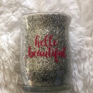 May include: A clear glass jar filled with silver glitter. The words "hello beautiful" are written in pink script across the front of the jar. The jar sits on a fluffy white surface.